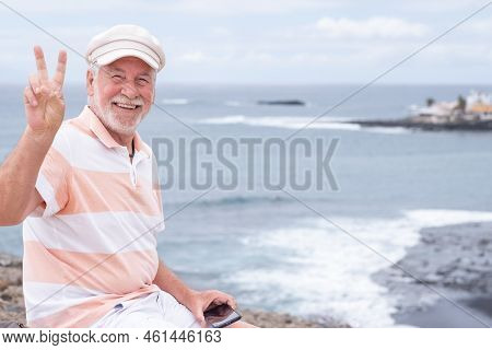 Smiling Beautiful Senior Man With Beard And Hat Sitting Outdoors At Sea Looking At Camera Enjoying F