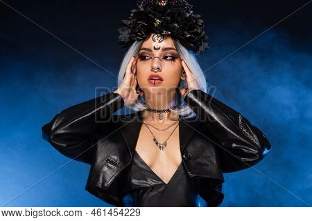Sexy Woman In Black Wreath And Witch Makeup Touching Ash Blonde Hair On Dark Background With Blue Sm