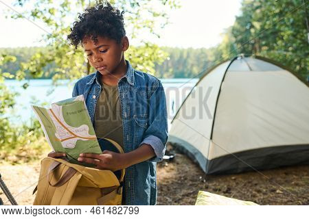 Cute Pre-teen Boy In Casualwear Looking At Map While Standing Against Tent