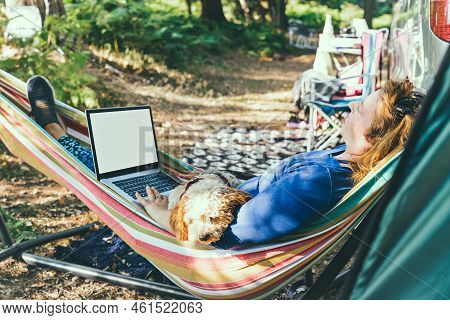 Adult Woman Working On Laptop Pc While Lying In Hammock, With Her Cockapoo Puppy Near Motorhome On C