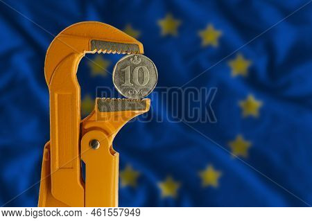 Kyrgyzstani Ten Som Coin Clamped In An Orange Plumber Wrench On The Background Of The European Flag.