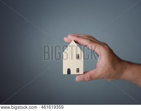 Concept Of Selling A House. A Hand Is Holding A Model House On A Gray Background. Real Estate Agent 