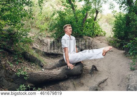 A Young Man Practices Yoga And Performs A Corner Stand On The Roots Of An Old Tree.