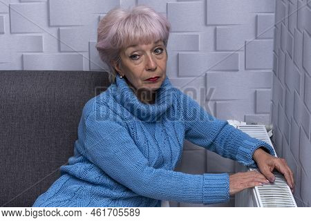 Elderly Woman 60-65 Years Old In A Warm Sweater Warming Near The Heating Radiator. Concept: Low Room
