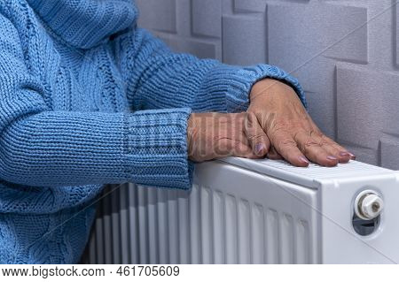 An Elderly Woman In A Blue Sweater Warming Her Hands On A Heating Radiator. Concept: Low Room Temper