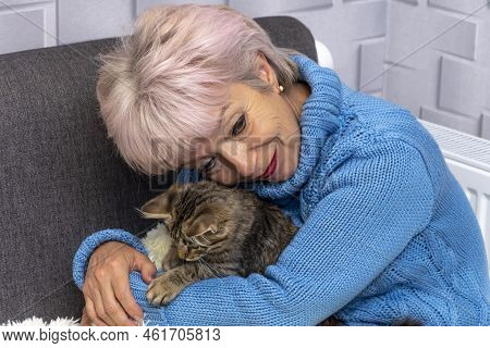 Elderly Woman 60-65 Years Old In A Warm Sweater Cuddling A Kitten, Sitting On The Bed By The Radiato