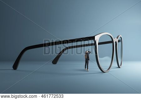 Thoughtful Young Businesswoman Looking Through Abstract Huge Glasses On Gray Background. Clear Busin