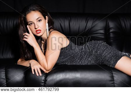 Elegant Woman In Lurex Dress And Shiny Earrings Lying On Leather Couch And Looking At Camera Isolate
