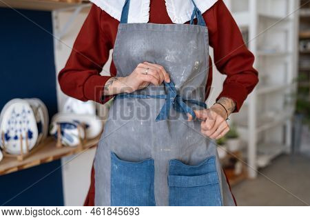 Female Ceramist Tying Her Apron Before Working With Raw Clay In Pottery Studio. Young Student Girl O