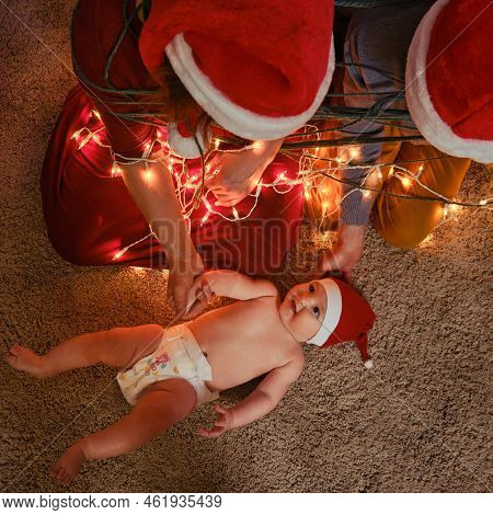 A Family With A Newborn Baby At The Christmas Tree In The Evening Living Room. Unfree Parents With A