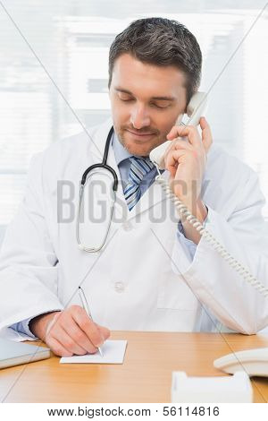 Concentrated male doctor using phone while writing notes at the medical office