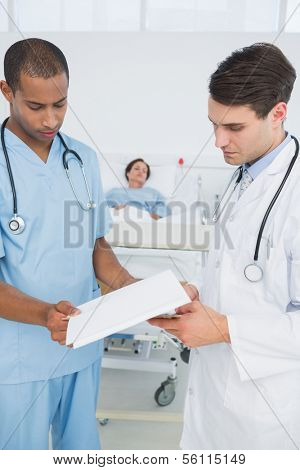 Doctor and surgeon looking at reports with patient in background at hospital