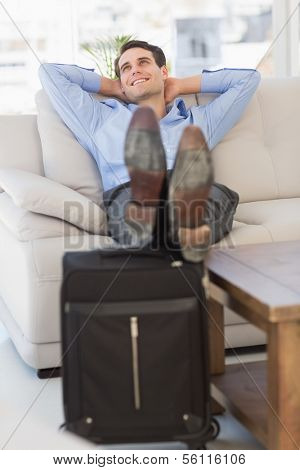 Smiling businessman relaxing on couch with feet up on suitcase in the office