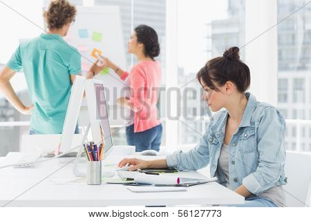 Side view of a casual woman using computer with group of colleagues behind in a bright office