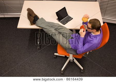 man sitting at with legs on the desk with hamburger and mug, heaving break for meal in office