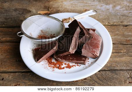 Bittersweet chocolate chunks with strainer of cocoa on white plate and rustic wooden planks background