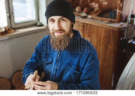 Worker with a beard wearing blue jeans suit and black hat carving wood and looking at camera, woodcarving instruments on table, portrait, copy space.