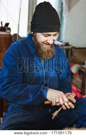 Man with a beard wearing blue jeans suit and black hat carving wood with instruments and looking at wooden spoon, smiling, portrait.
