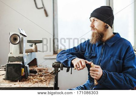 Man with a beard wearing blue jeans suit and black hat sitting on working place with wooden spoon and looking left, woodcarving instruments on table.