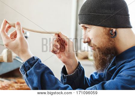 Worker with a beard wearing blue jeans suit and black hat holding and looking at wooden spoon, woodcarving, portrait, copy space, close up.