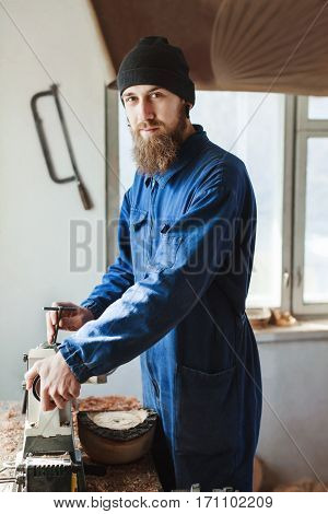 Worker with a beard wearing blue jeans suit and black hat working near table, woodcarving instruments on table, big window at background, portrait.