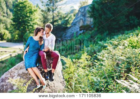 Couple sitting on the small rock, outdoor. Spectacular view, nature, many trees and fern. Sunny day, summer, blue sky.  Girl wearing blue dress and light blue shoes and man wearing white shirt and claret trousers. Full body
