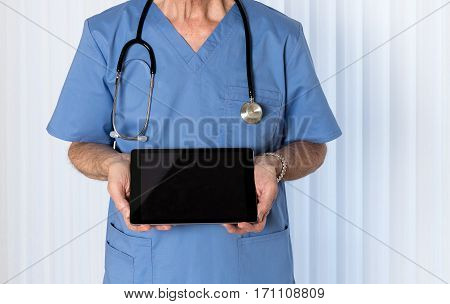 Senior male caucasian doctor with stethoscope in medical scrubs looking up and holding electronic tablet for message