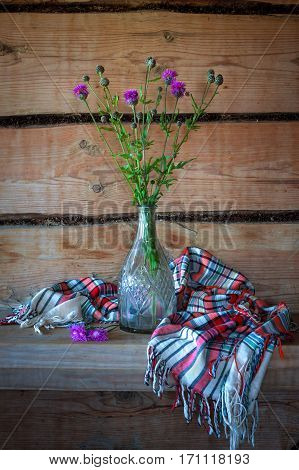 Beautiful still life with purple flowers in a glass vase and a beautiful shawl in the old rural wooden house