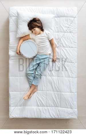 Better than Ringo. Cute committed charming child falling asleep in his bed after playing drum and still holding in in his hands