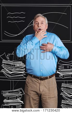 Heavy workload. Nice aging stressed man standing among piles of paper and holding his hand near his heard while working at the office