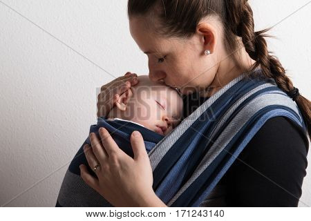 Beautiful young mother with her baby son sleeping in sling, kissing him on head. Studio shot on white background.