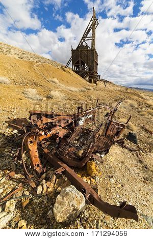 Ghost town mining head frame in the Nevada desert with wrecked car in the foreground. Sky is blue with clouds.