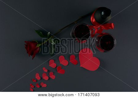 Wine bottle and two wineglasses with red hearts and rose flower on a dark background. Love card concept Valentine's day theme. Shot from above