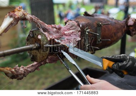 Barbecue picnic at country fair. Lamb roasted at spit. Grill, big piece of meat at rolling skewer. Street vendor, professional chef hands cooking bbq food, unrecognizable. Catering concept