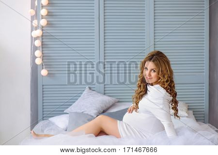 young beautiful pregnant woman with curly hair and white dress lying in bed and smilling
