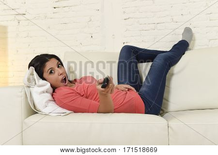 young beautiful Spanish woman in jeans home alone watching television laughing lying at sofa couch on living room happy and excited enjoying comedy movie having fun holding remote