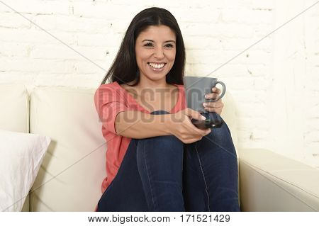 young beautiful Spanish woman in jeans home alone watching television smiling sitting at sofa couch on living room happy and excited enjoying TV romantic comedy movie relaxed