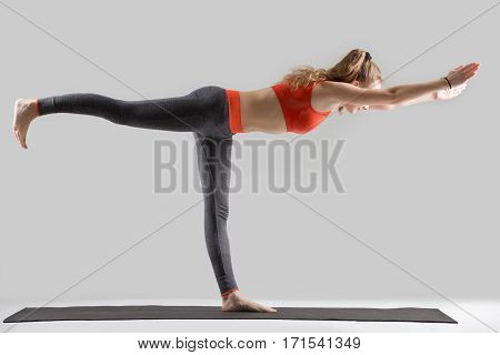 Young attractive woman practicing yoga, standing in Warrior Three exercise, Virabhadrasana III pose, working out, wearing sportswear, red sports bra, pants, indoor full length, isolated, grey studio