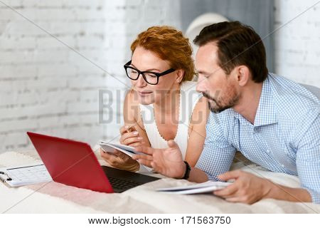 Listen to my idea. Optimistic involved delighted couple lying on the bed at home and using the laptop while sharing points of view and taking notes