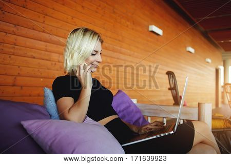 Young hipster girl is talking via mobile phone with her friend during work on portable net-book while is sitting on hotel terrace. Smiling pretty woman having pleasant cell telephone conversation