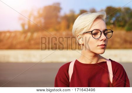 Stylish female hipster with short haircut and trendy eyewear traveling alone on sunny autumn day resting on urban setting blurred background near copy space for your advertising content or text