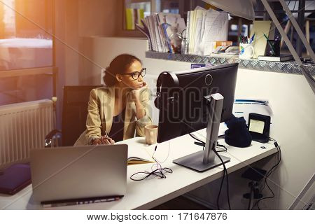 Professional female newsmaker monitoring latest event report from official media sites via modern laptop computer connected to wireless network working hard at night drinking coffee sitting in office