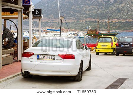 Bali Greece - April 30 2016: White sport-car Audi A5 coupe parked on Mithos beach promenade in sea bay of resort village Bali. Classic Greek stone architecture of resort tourists walk streets of city. Rethymno Crete Greece
