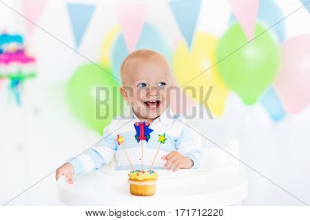 Adorable baby boy celebrating first birthday blowing candles on colorful cup cake. Kids birthday party decorated with balloons and pastel color banner. Child eating cake and candy.
