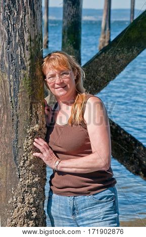 This portrait is a natural looking middle aged Caucasian woman in her fifties. She's wearing glasses blue jeans and a brown tank top and leaning against some ocean pillings with barnacles attached.
