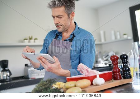 Mature man in kitchen cooking with help of receipe on tablet