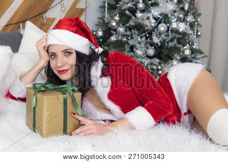 Pretty Woman In Santa Dresses With Gift Lie Prone On The Bed In A Room On The Background Of The Chri