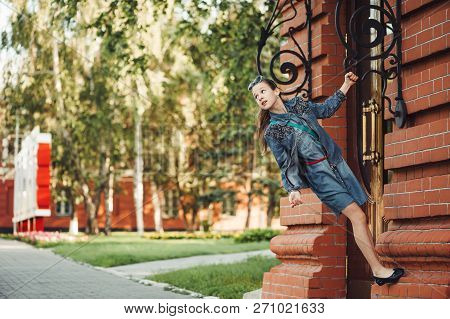 Young Cheerful Child Piddle Near Door Of Building From Red (brown) Brick Near Green Grass Outdoors. 