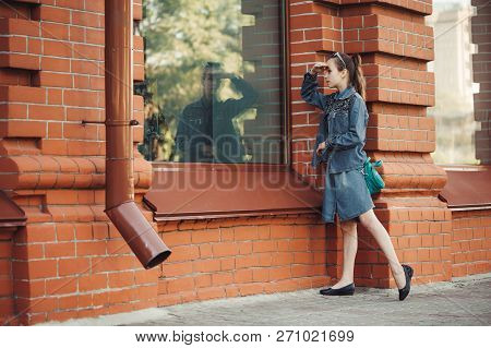 Teenage Girl In Blue Denim Dress Look In Window Of Building From Red (brown) Brick Outdoors. Young S