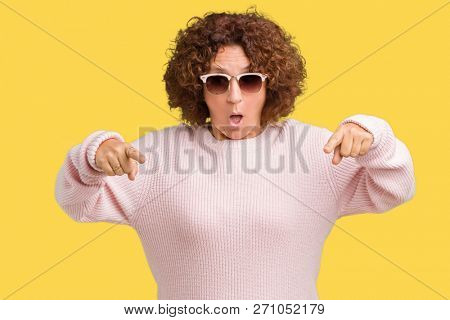 Beautiful middle ager senior woman wearing pink sweater and sunglasses over isolated background Pointing down with fingers showing advertisement, surprised face and open mouth